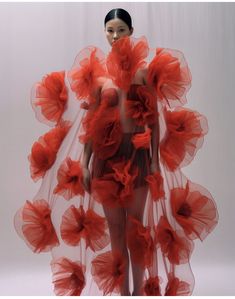 a woman in a red flowershaped dress with seethrough mesh fabric. she is standing in front of a white background. the dress is made up of many individual flower shapes, which are all connected together. the flowers are all made of a soft, delicate material. she looks fashionable and elegant.