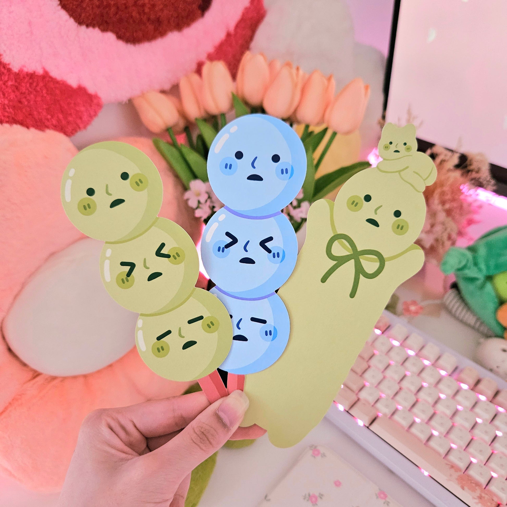 the image shows a hand holding three adorable illustrated bookmarks. two bookmarks are shaped like dango (japanese sweet dumplings)  one in light green and the other in light blue. each dango has a cute, slightly sad face.  the third bookmark is shaped like a cute, pale green ghostlike character with a bow.  all characters have simplified, expressive eyes and mouths.  the background includes a pink fluffy object, a bouquet of pastel tulips, and a pink keyboard, creating a very soft and aesthetically pleasing scene. the overall style is kawaii and pasteltoned.