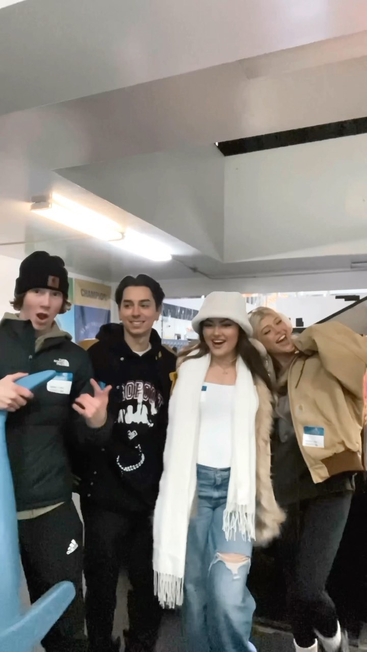 a group of four young adults posing indoors. the first person is a young man with reddishbrown hair wearing a black beanie, a dark green puffer jacket, and black pants. he's holding a blue toy and has a name tag on his chest. next to him is a young man wearing a black sweatshirt. the third person is a young woman wearing a white bucket hat, a white scarf, a white top, and ripped blue jeans. the fourth person is a young woman with blonde hair wearing a tan jacket and black jeans. she also has a name tag on her chest. they all appear to be smiling and having a good time. the background includes a white ceiling with lights and some signage.