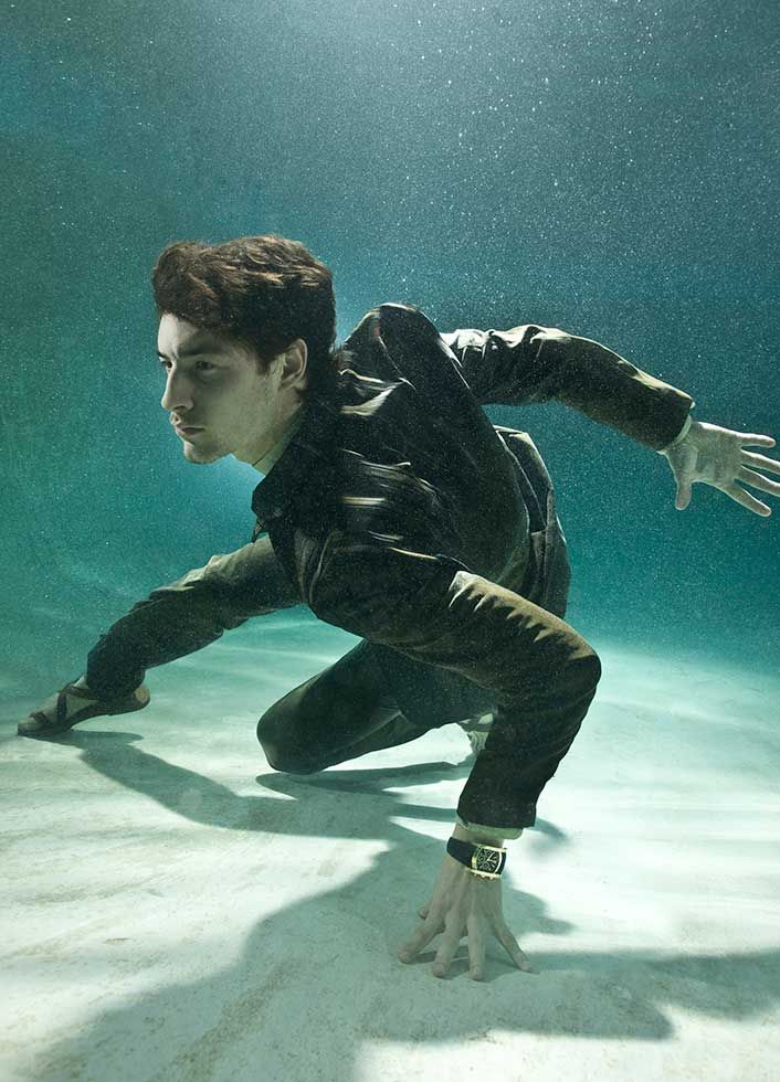 a man in a suit is underwater, diving and swimming in a pool. he is wearing a luxury gold watch. he looks fashionable and elegant, as if he's trying to keep his composure while gracefully swimming.