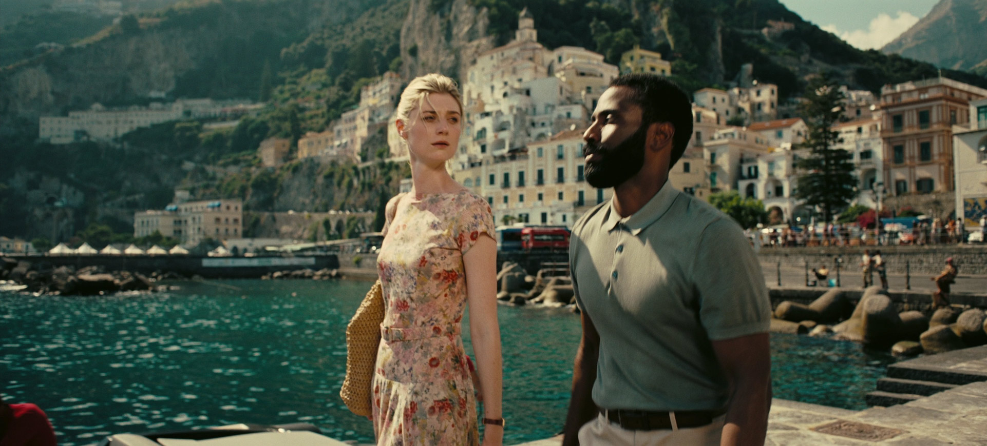 a man and a woman stand together on a dock in positano, italy, with the picturesque town rising behind them and the sparkling mediterranean sea in the background. the woman, positioned slightly ahead of the man, gazes towards the town while he looks straight ahead.  the image evokes a sense of romance, travel, and the beauty of the italian coast.