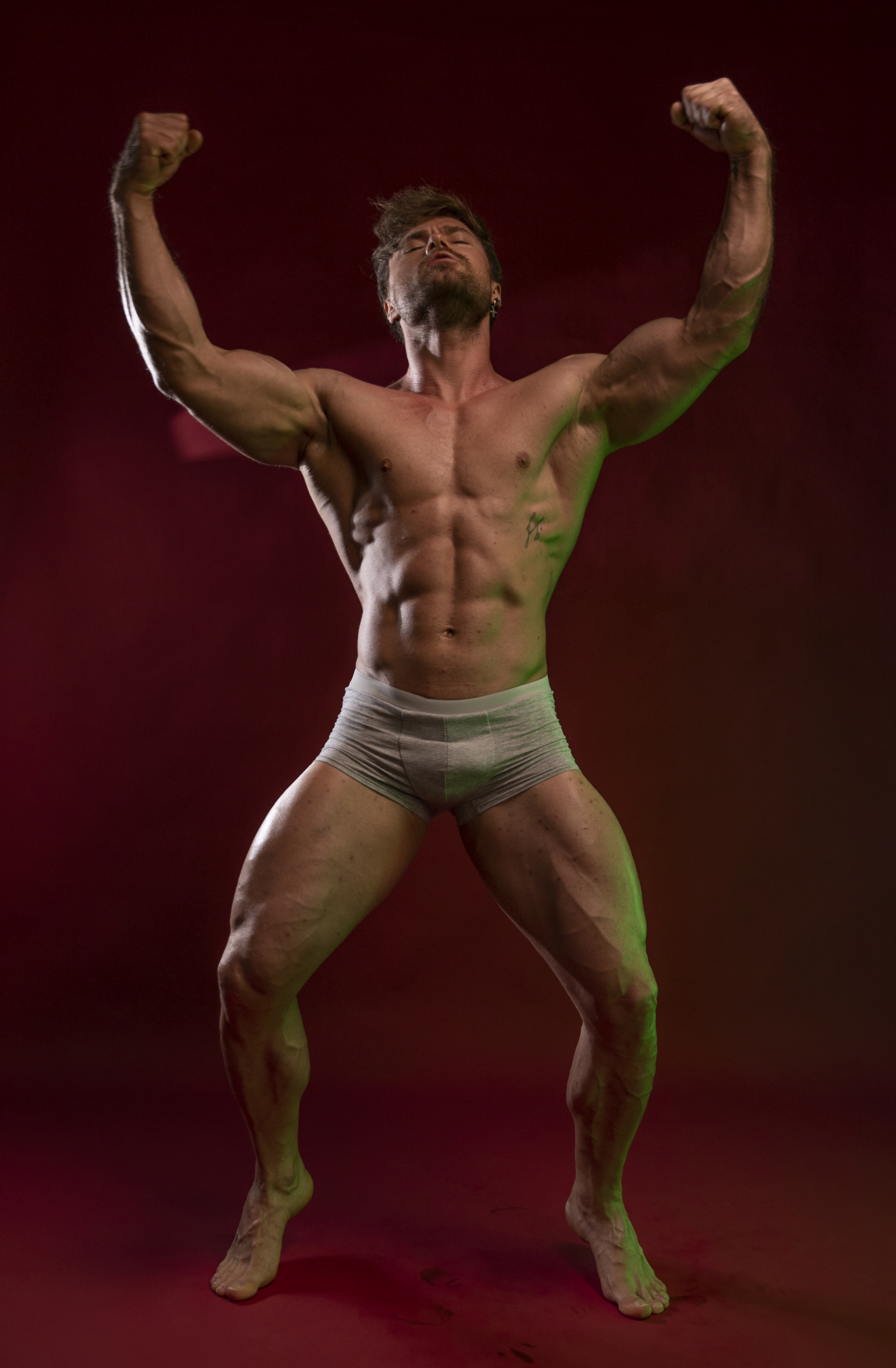 a muscular, shirtless man is flexing his biceps and abdomen in a studio shot. he is wearing underwear and his body shows the results of fitness training. he is standing against a dark red background with studio lighting.