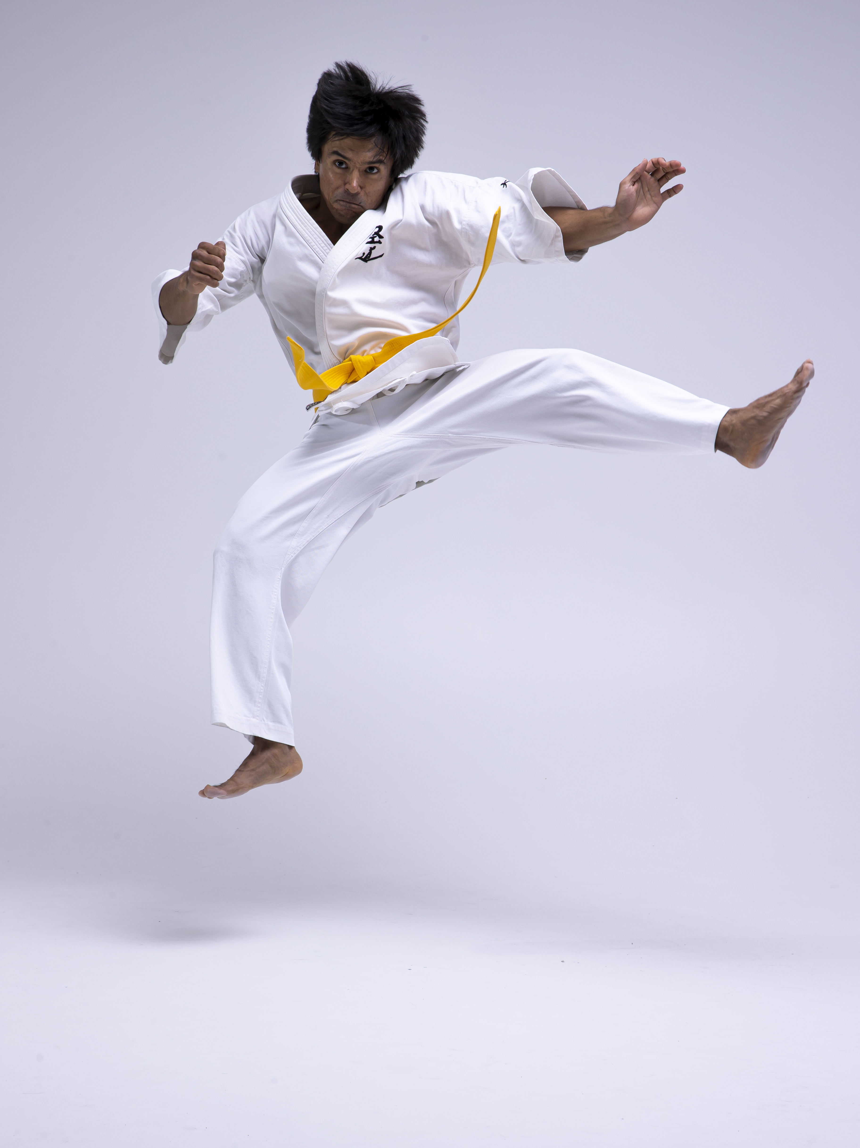 a man wearing a white karate gi with a yellow belt is jumping in the air. he is barefoot and has one leg extended straight out. he appears to be performing a karate kick. the background is white.