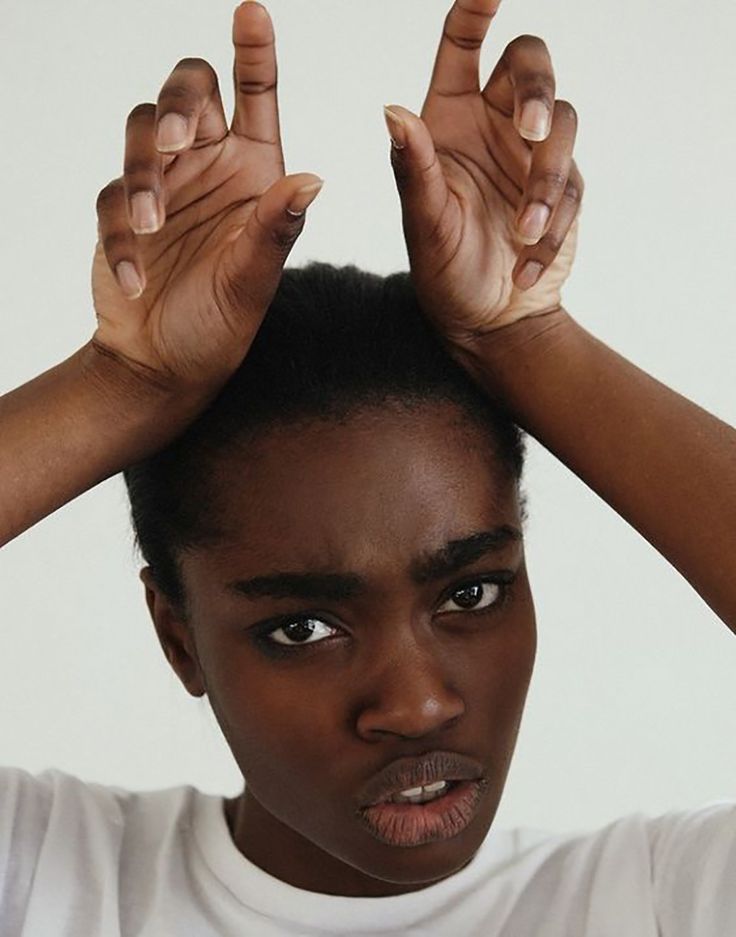a young black woman in a white shirt with a serious expression on her face. her arms are raised, her hands are forming a 'v' shape, and her hair is black. she is looking directly at the camera.