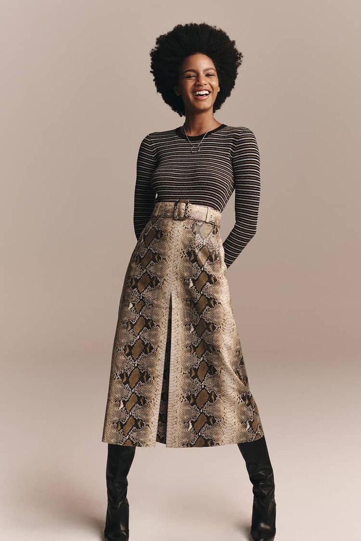 a black woman with an afro is smiling. she is wearing a black and white striped shirt, a snake print skirt, and black boots. she is posing for a photo.