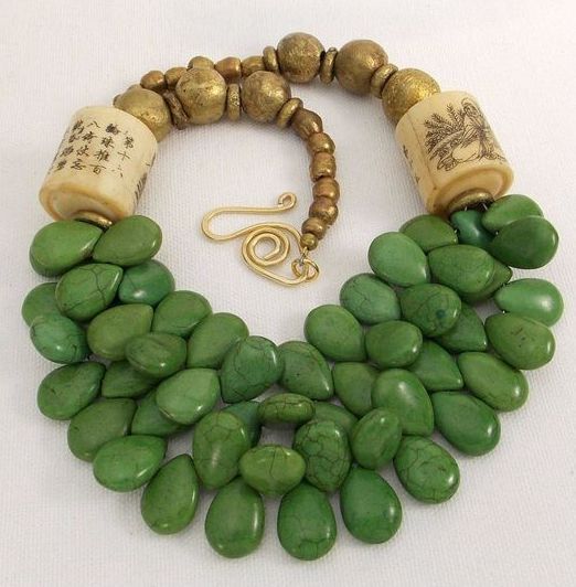 the image shows a necklace featuring a combination of green stones, gold beads, and cylindrical beads with inscribed details. the green stones are arranged in a layered, teardrop shape, creating a biblike effect. above the stones, there are gold beads in varying sizes, leading to the cylindrical beads, which appear to have writings or drawings on them. the necklace is fastened by a gold clasp with a spiral design. the piece appears to be handcrafted and artistic.