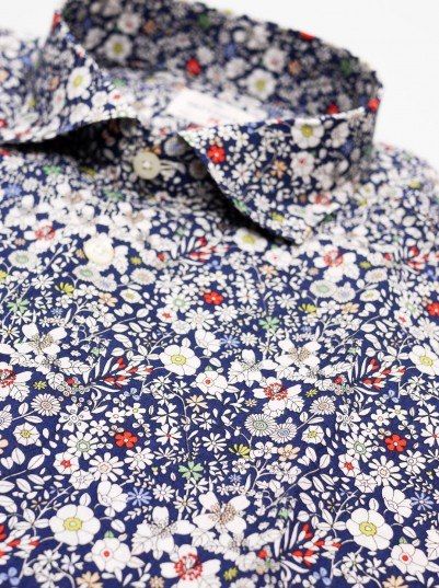 the image shows a closeup view of a men's buttondown shirt. the shirt is made of a navy blue fabric with a dense, allover liberty print featuring a multitude of small, multicolored flowers and leaves. the collar is visible, showcasing the same detailed floral pattern. the focus is on the intricate and delicate design of the fabric, highlighting its texture and the variety of floral motifs. the image gives a strong impression of a highquality, potentially premium, garment.