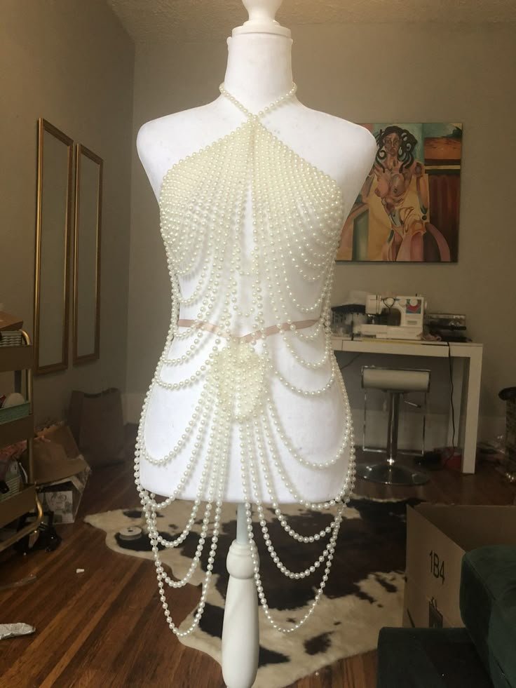 a white mannequin wearing a dress made of pearls, on a wooden floor with a cowprint rug. the dress is a halter top style with a necklace strap and draped pearl strands forming the bodice and skirt. in the background, there is a painting on the wall, a desk with sewing equipment, and a cardboard box.