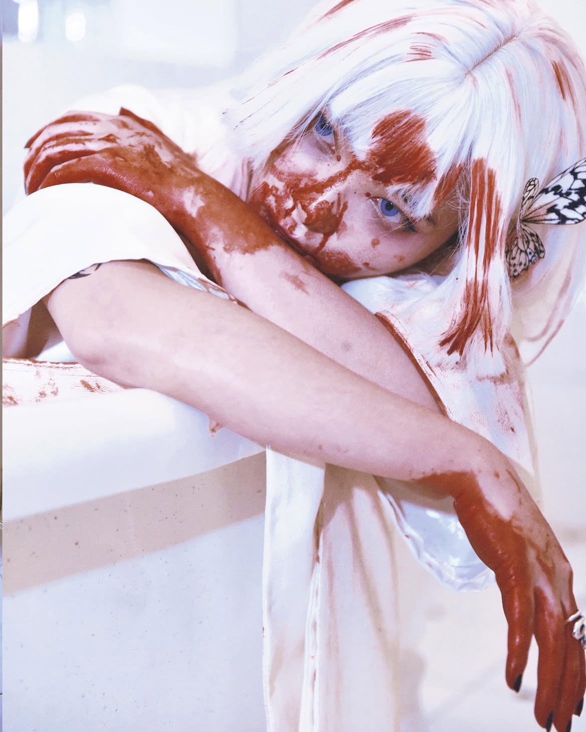 a woman with white hair and blue eyes is shown covered in what appears to be blood. she is wearing a white dress and has a butterfly attached to her hair. her expression suggests sadness or distress. she is wearing a ring on her finger. the makeup gives her an artistic look, rather than one that feels real.