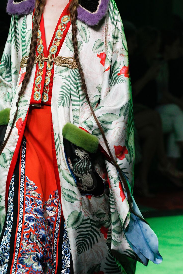loseup detail shot of a model showcasing a gucci outfit on a runway. the garment is a long, flowing coat or robe featuring a predominantly white background with a vibrant floral print incorporating green ferns and red poppies.  the coat has long sleeves, a fur trim at the collar and cuffs, and an embroidered, ornate detailing along the front placket and hem.  a red underdress peeks out from beneath. the model's long braided hair is visible. the backdrop suggests a runway setting.  the overall aesthetic is luxurious and elegant.