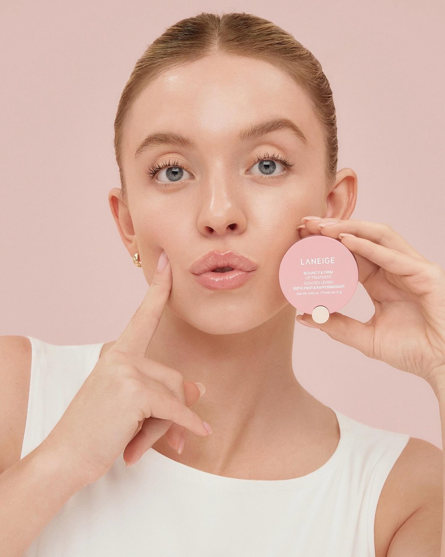 closeup shot of a young woman with flawless skin holding a laneige cushion compact.  she's touching her cheek with one hand and holding the compact with the other. her skin appears radiant, dewy, and healthy, reflecting the product's promise. the background is a soft pink, complementing the product packaging and the overall aesthetic. the image focuses on the woman's glowing skin and the product in her hands, emphasizing beauty and skincare.
