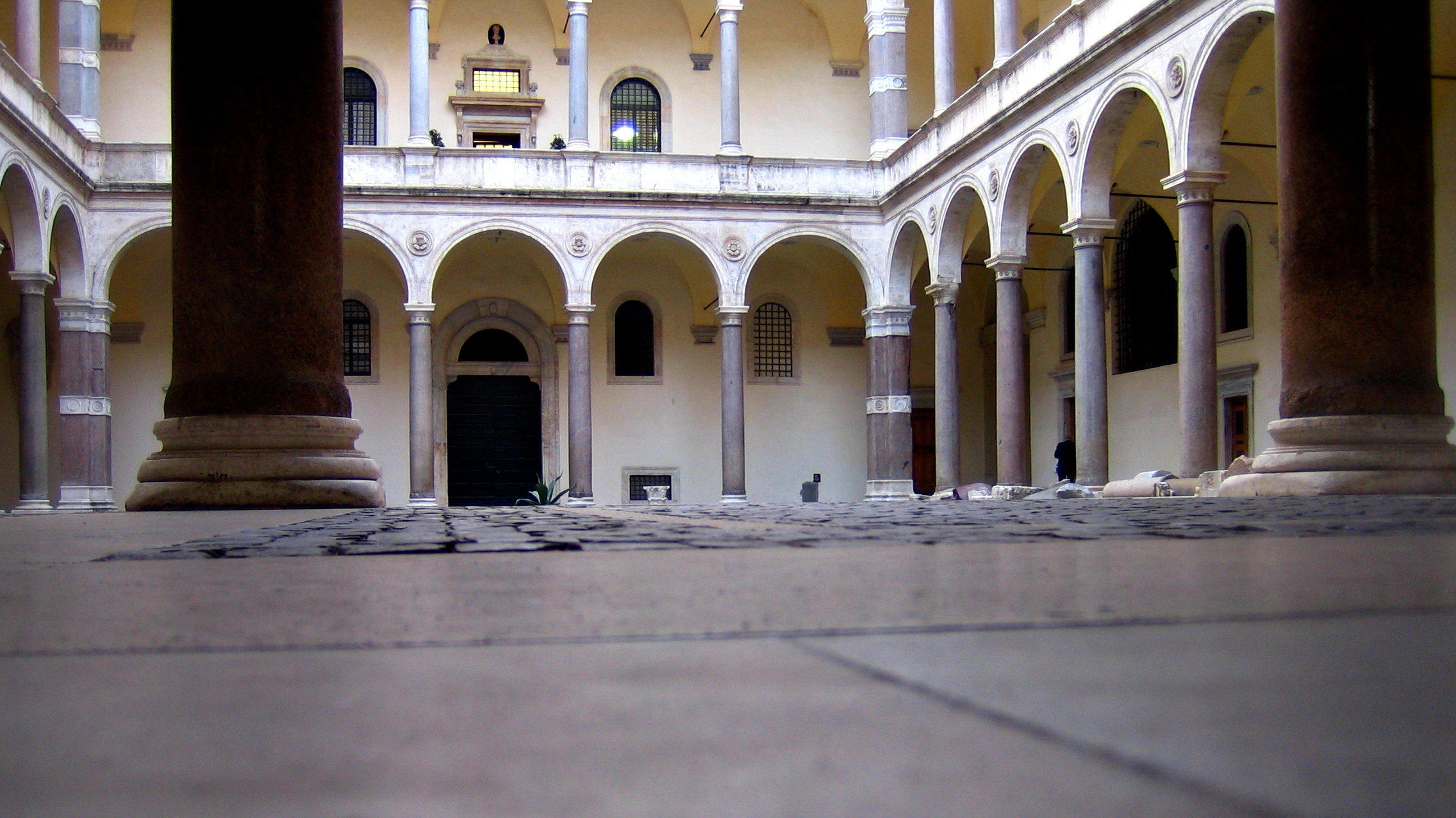this is a lowangle shot of a courtyard surrounded by a twostory building with a classical design. arches supported by columns line the perimeter of the courtyard, and there are large, brown, cylindrical columns that frame the view from the foreground. there are windows and doors visible behind the arches, and the courtyard is paved with stone.