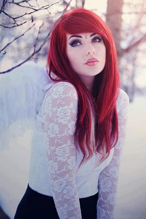 a young woman with vibrant red hair and pale skin poses for a portrait against a snowy backdrop. she wears a delicate white lace top, and white feathery angel wings peek out from behind her. her makeup features dramatic eyeliner and a pale pink lip. the overall feel is ethereal and whimsical.