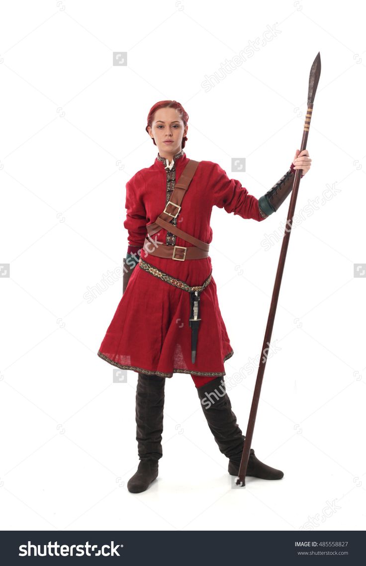 a woman dressed in a red medieval costume, stands with her right foot raised in the air,  with a spear held in her left hand. she is looking directly at the camera.