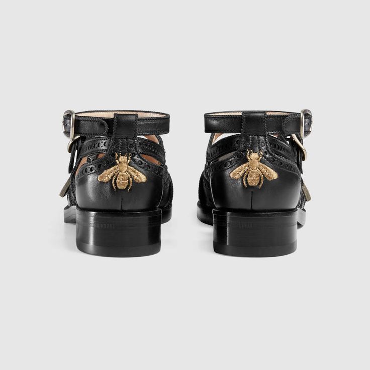 a pair of black gucci women's leather shoes are shown from the back. the shoes are ankle strap shoes with brogue detailing and bee embroidery.  they have cutouts and perforated patterns. the shoes are positioned sidebyside against a light gray background.