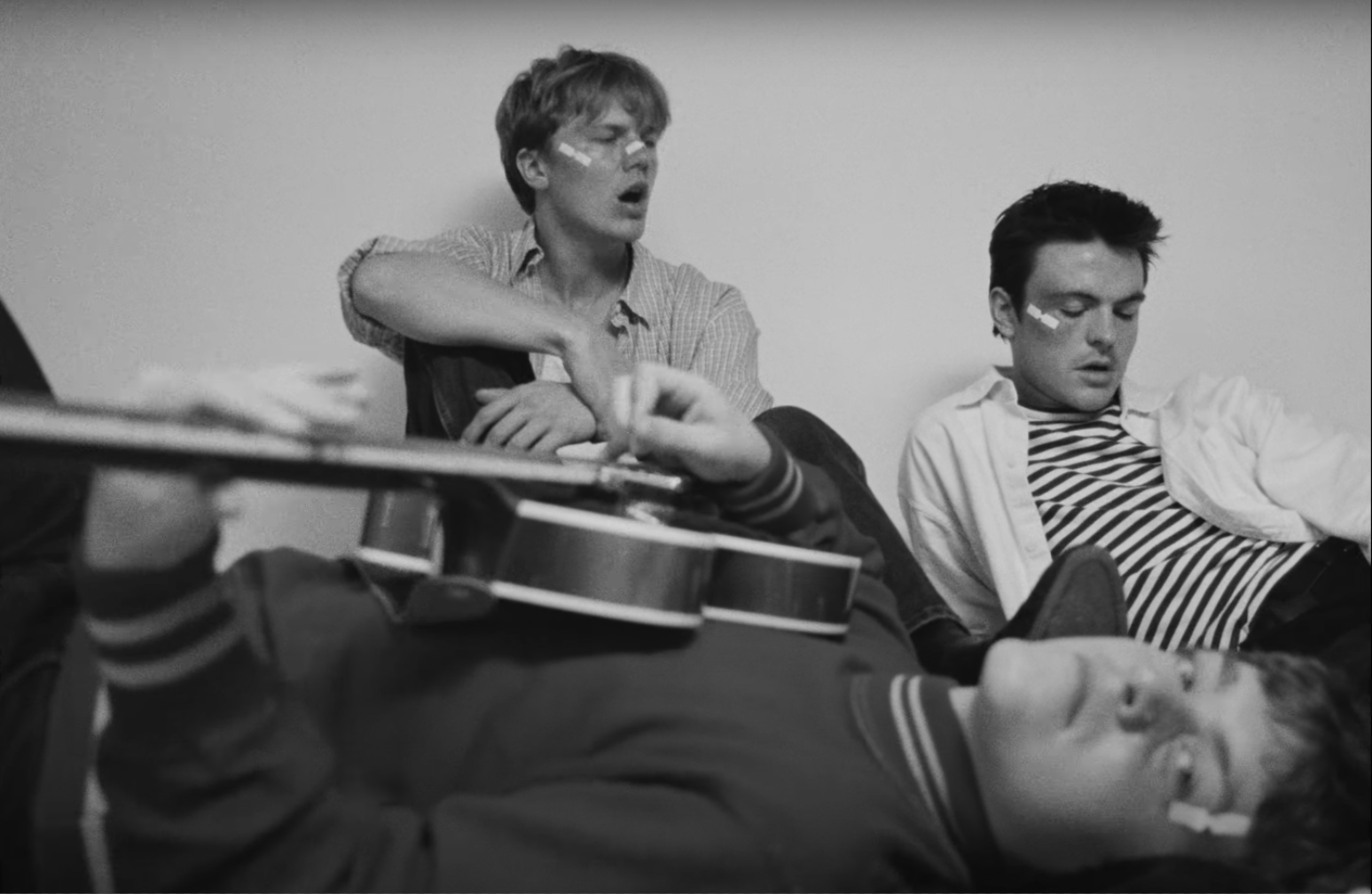 three young men are sitting together in what looks like a dorm room. one is laying down with his guitar, while the other two are sitting and looking off into the distance. all of them have white tape on their faces.