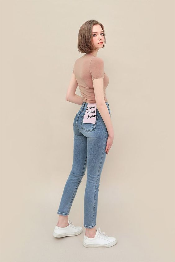 a young woman poses against a beige backdrop, showcasing a stylish and casual outfit. she wears a fitted, light brown tshirt tucked into a pair of highwaisted skinny jeans. the jeans have a unique label on the back pocket reading "chuu 5kg jean." the woman stands with her back to the camera, glancing over her shoulder with a neutral expression. she completes the look with simple white sneakers. the soft lighting and neutral background create a clean and minimalist aesthetic, highlighting the outfit and the model's pose.