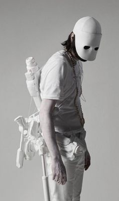 a person in allwhite clothing and a white mask. they are standing in front of a light grey background. the person is wearing what appears to be some type of white sculpture or device on their back. the clothing and the mask are both very minimalist and futuristic in style. it appears to be a piece of art and is very surreal and interesting.