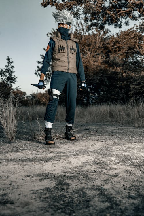the image shows a person in a detailed kakashi hatake cosplay from the anime/manga series naruto.  they are standing outdoors in a barren, dusty landscape under some trees. the cosplayer is wearing a beige vest over a dark blue shirt and pants, along with kakashi's signature headband and sandals. they are holding a kunai (throwing knife) and have a serious facial expression. the overall setting and costume replicate the look and feel of the naruto universe.