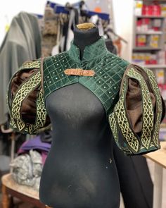 a closeup shot of a mannequin dressed in a green and brown medievalstyle vest. the vest is made of velvet and features intricate embroidery on the sleeves. the mannequin is positioned in a retail store setting.
