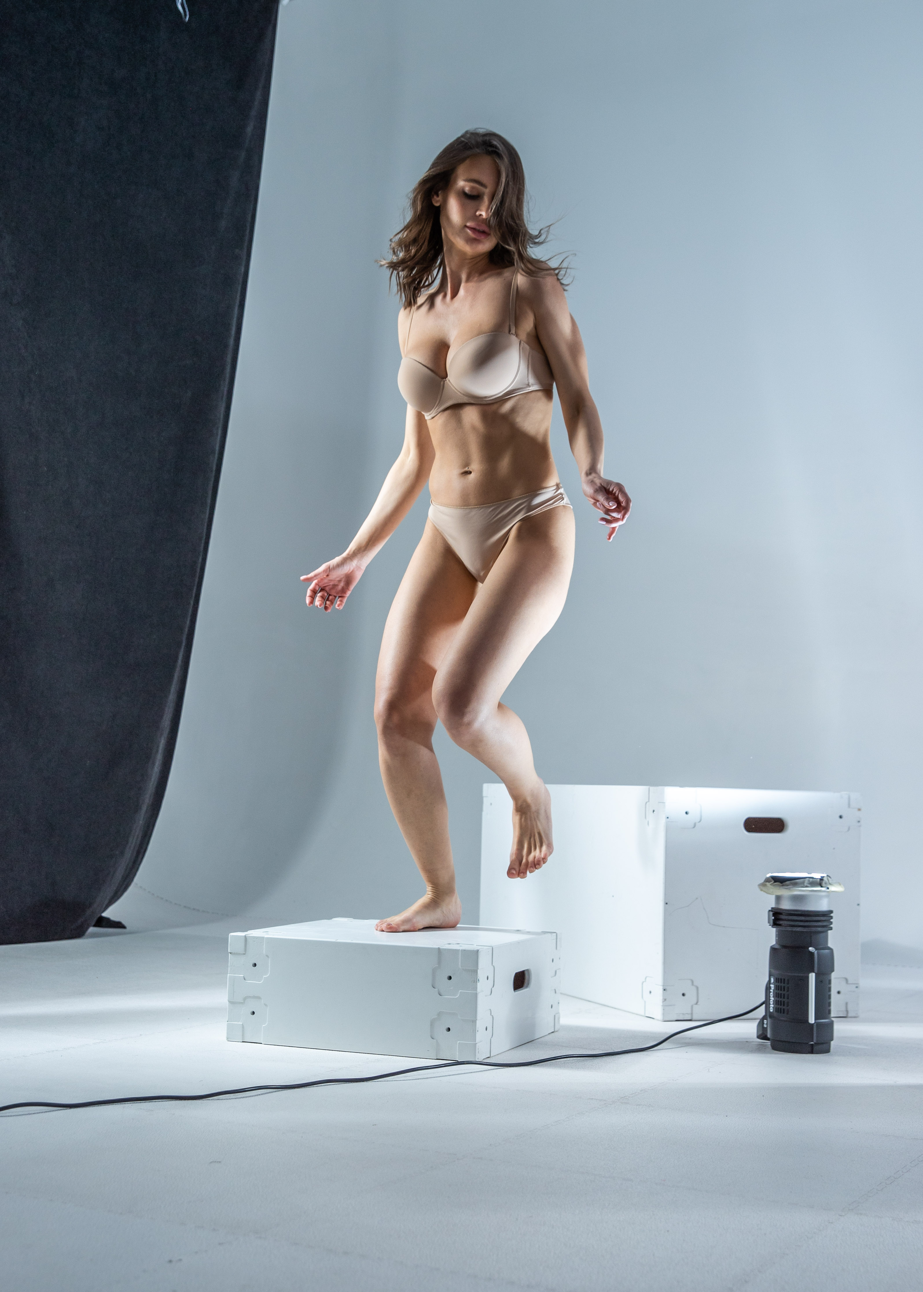 a woman wearing nude lingerie is jumping off a white box in a studio. she is barefoot and her hair is down. there is a gray backdrop behind her, and another white box and a light are next to the box she is jumping from.