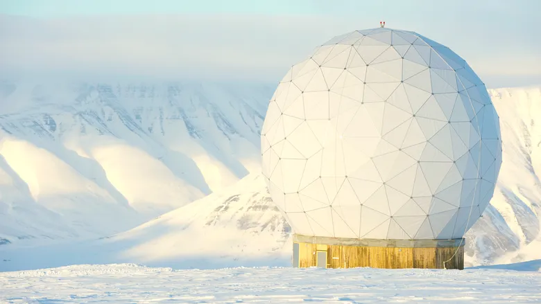 a large white geodesic dome, resembling a radar antenna, stands against a snowy mountainous backdrop. the dome is perched atop a wooden platform, with the surrounding landscape blanketed in pristine snow.  the scene evokes a sense of isolation, scientific research, and a remote outpost.  the cold, clear air and stark beauty of the arctic landscape create a dramatic contrast to the futuristic structure.