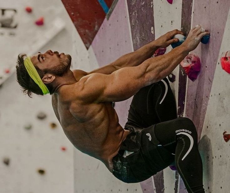 a man wearing black compression pants and a green headband is rock climbing indoors, he is using his grip to hold onto the wall.