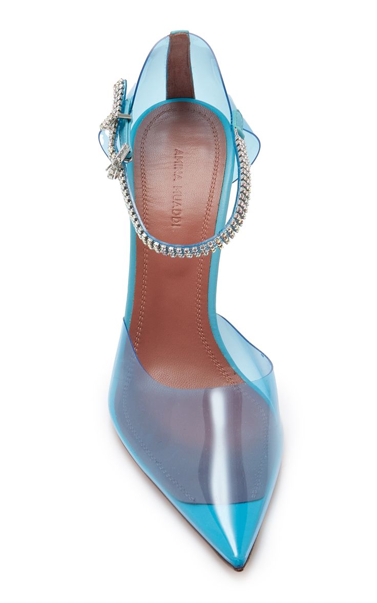 pair of amina muaddi blue transparent pvc high heels are shown in the image.  the shoes have a pointed toe, a crystalembellished ankle strap and a light brown insole.  the shoes are angled slightly toward the camera showing the details of the upper and sole.  the background is white.