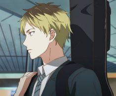 this is an anime style drawing of a young man from his shoulders up. he has short, blond hair and an earring. he is wearing a white button down shirt with a blue tie, and a navy blue suit jacket. he is carrying a large black instrument case on his back. he is standing sideways with his head slightly turned. the background is blurred.