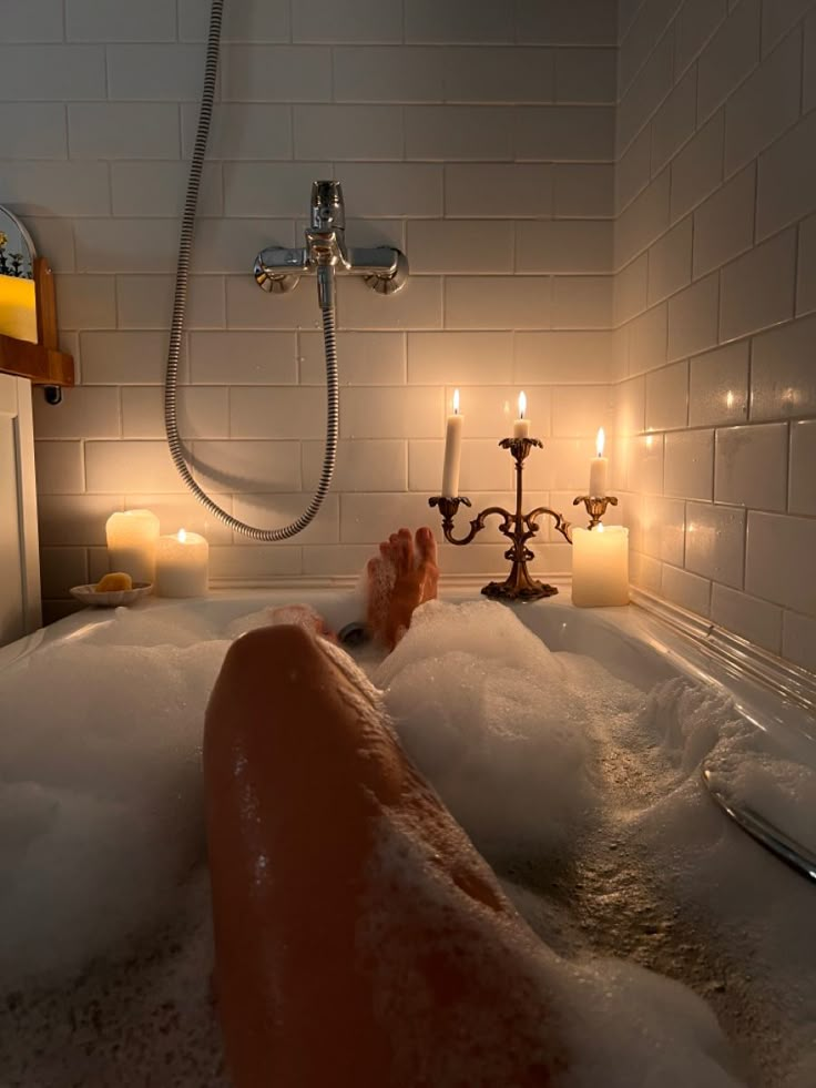 a person is relaxing in a bubble bath, with candles lit around the tub. the bathroom has white tiled walls and a chrome shower head. the scene creates a warm and relaxing atmosphere.
