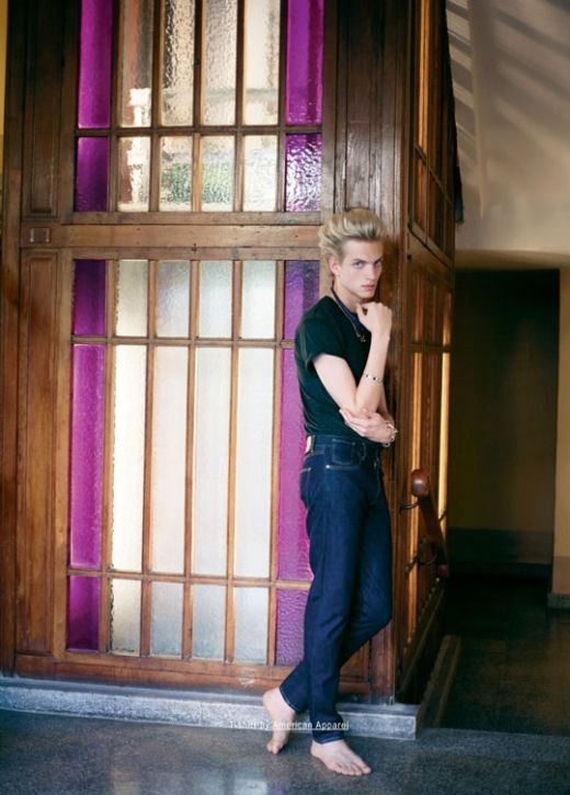 a young, attractive male model with blonde hair stands barefoot in a hallway, leaning against a wooden door with purple stainedglass panes. he's wearing a darkcolored shirt and darkwash jeans. the photo is a fashion portrait, highlighting the model's style and the clothing. the setting is an indoor location, likely a building with an oldfashioned aesthetic.