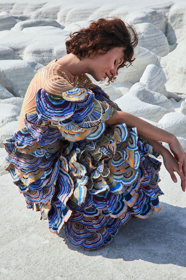 a woman with curly hair sits on white travertine terraces, wearing a unique dress with blue, orange, and brown layered patterns. she is looking down with a thoughtful expression. the background features more of the distinctive white rock formations.