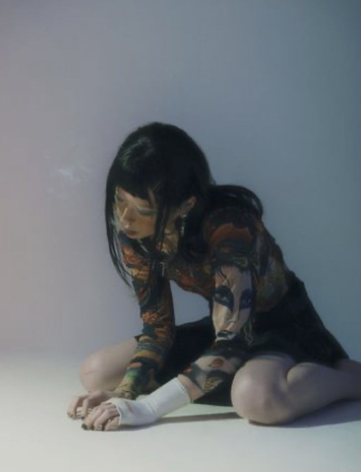a young woman with black hair is sitting on the floor in a studio. she is wearing a black longsleeved shirt with colorful tattoos and black pants. she has a white bandage on her left arm. she is looking down at her hands and is resting her head on her left hand.