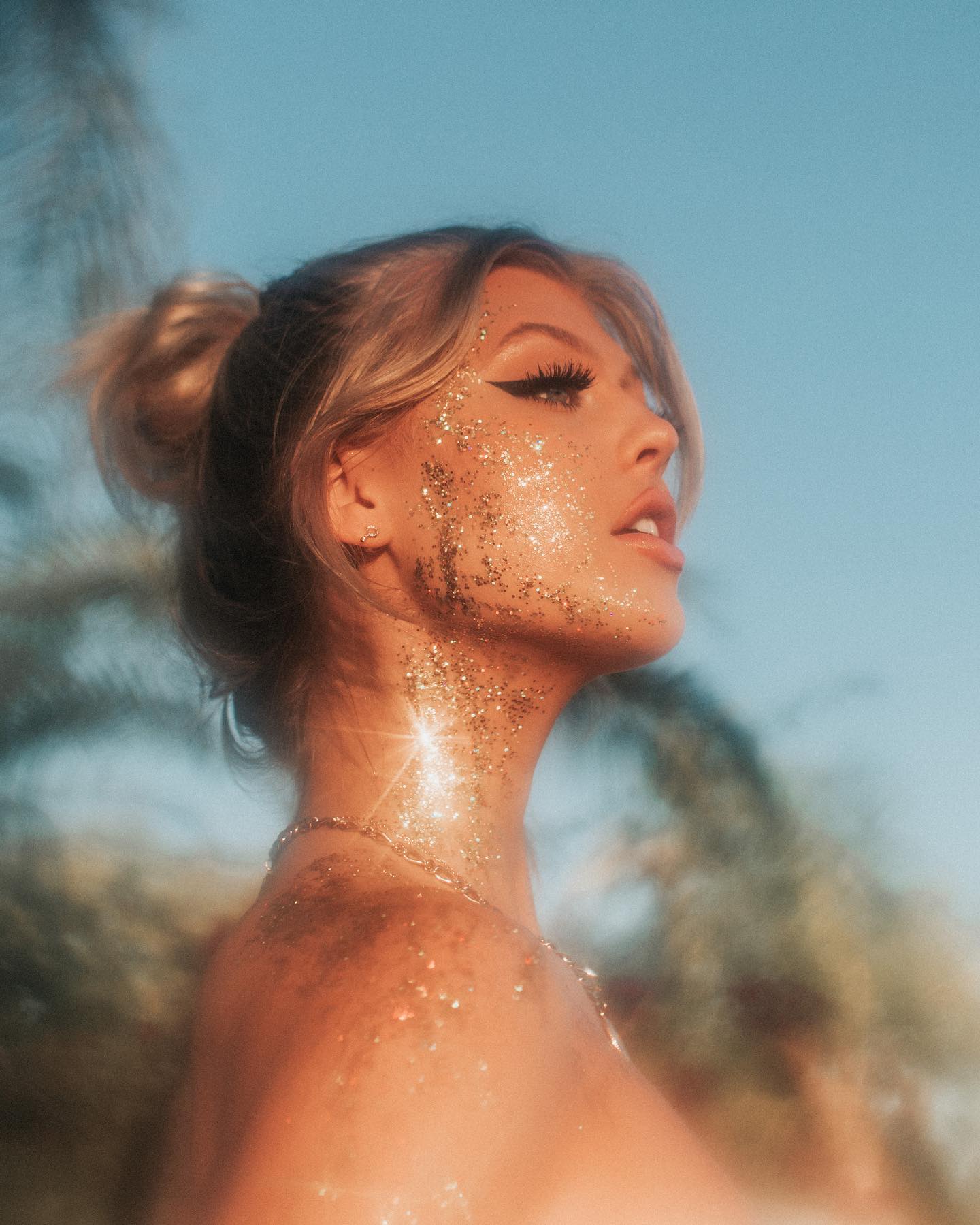 a woman with blonde hair pulled back in a bun, wearing glitter makeup on her face and neck, and a gold necklace, stands outdoors with a blue sky and palm trees in the background. she is looking up and to the right, and sunlight is shining on her skin. her shoulder and collarbone are visible.