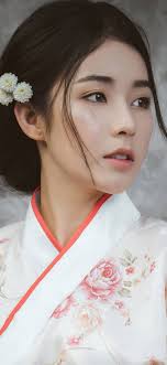 a young asian woman, dressed in a white and pink floral kimono, is the focal point of this portrait. she has dark hair styled in an updo with small, white flowers adorning it. her makeup is minimal, highlighting her natural beauty. the background is blurred, creating a sense of intimacy and focusing the viewer's attention on the woman's delicate features and serene expression.
