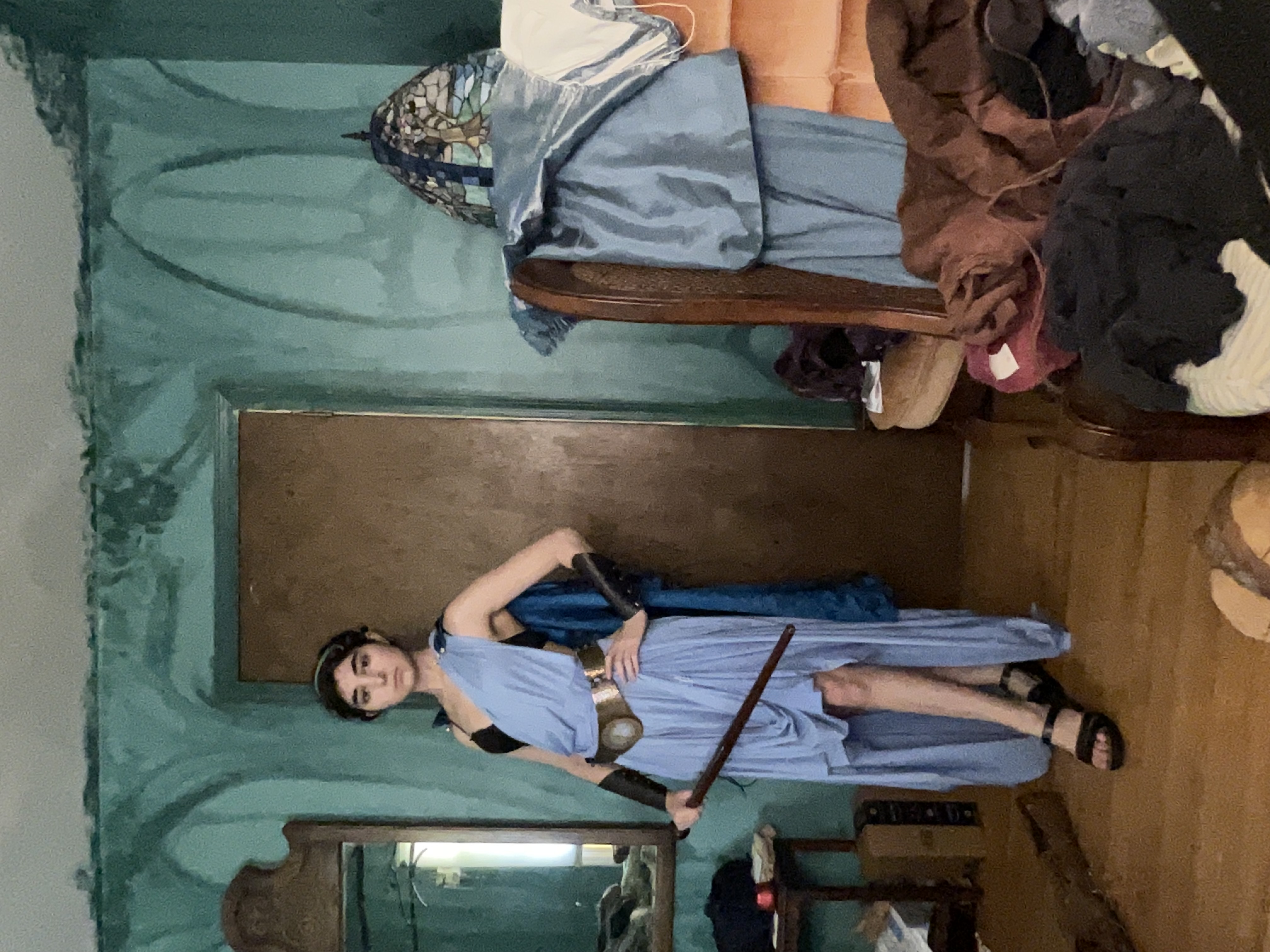 a young woman in a homemade cosplay outfit stands in a cluttered room.  she's dressed as a greek warrior goddess, wearing a light blue highlow dress, brown sandals, a gold belt, and dark armbands. she holds a long, dark sword. her hair is styled up, and she wears a headband. the room's walls are a teal green, and various items are piled on a chair in the background. an antique mirror is visible in the left corner.  the overall aesthetic is a mix of fantasy and a slightly messy, livedin space.