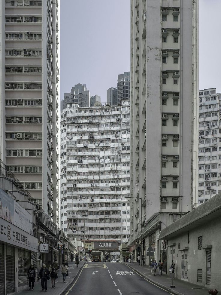 a monochrome photograph depicts a street scene in hong kong.  tall, densely packed apartment buildings line both sides of a relatively narrow street.  a building in the middle distance appears older and slightly different in design than those on either side.  pedestrians are visible, walking along the sidewalks, adding a sense of scale.  the overall mood is quiet and slightly muted in color, emphasizing the architectural features of the city.