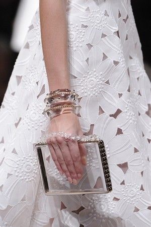 a woman in a white floral lace dress is holding a clear clutch. the dress is very delicate and feminine, with a lot of intricate detailing. the woman is wearing several bracelets on her wrist. she has her hand on the clutch, and the light is reflecting off the clear material.  the image is very feminine and elegant. it captures the beauty of a summer day.