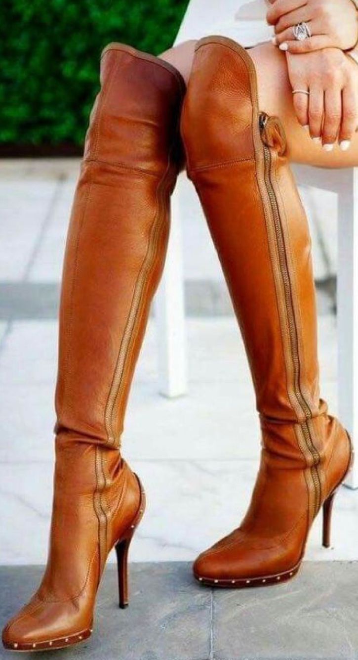 a woman wearing brown kneehigh boots with a high stiletto heel. she is standing with her legs crossed and her arms resting on her legs. the boots are made of leather and have a zipper up the back. she is wearing a white top and her skin is tanned. the image is taken from the waist down and the focus is on the boots.