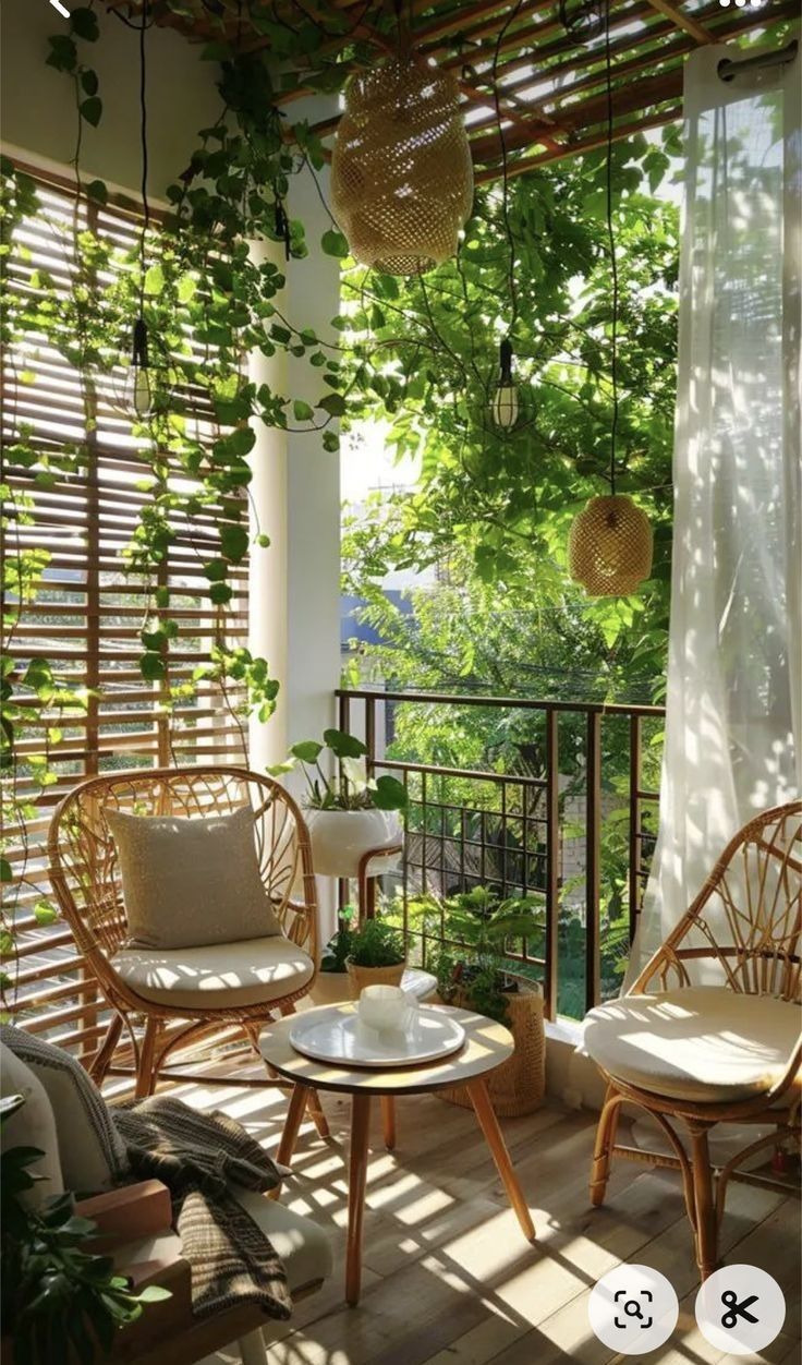 he image shows a cozy and inviting balcony setting. two rattan chairs with white cushions face each other around a small wooden coffee table.  the balcony is adorned with lush greenery, including climbing plants growing up a bamboo screen. hanging woven lights provide soft illumination, and sheer curtains add a touch of elegance. the overall atmosphere is relaxing and bohemian, with a tropical feel. sunlight streams in, highlighting the natural materials and the abundance of plants.