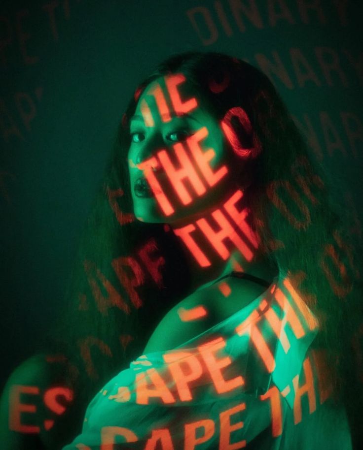a woman with dark hair and a green shirt is illuminated by a red light projection, casting words on her face and body. the background is a dark green, making the red words stand out. the overall mood is mysterious and cool.
