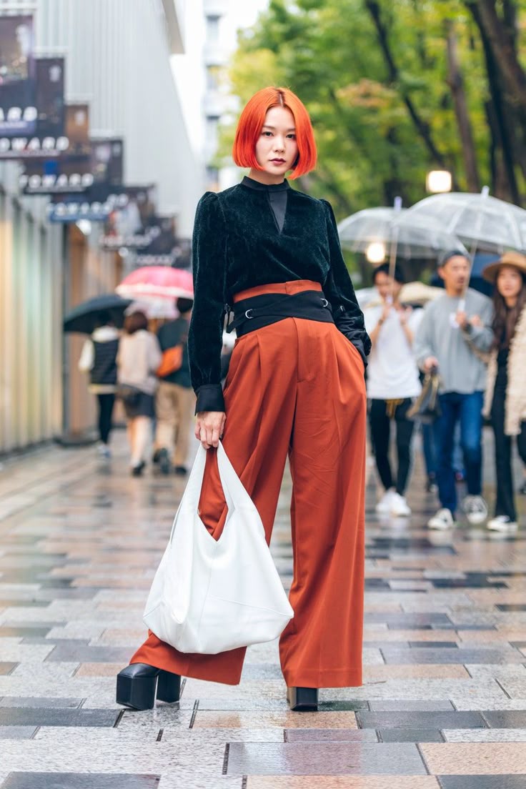 a stylish woman with short orange hair stands confidently on a wet paved street. she's wearing a black velvet longsleeved top, highwaisted, wideleg rustcolored pants with a wide black belt, and platform shoes. she's carrying a white shopping bag. behind her, people are walking with umbrellas on a rainy day. buildings line the street, and the overall ambiance is that of a fashionable urban setting.