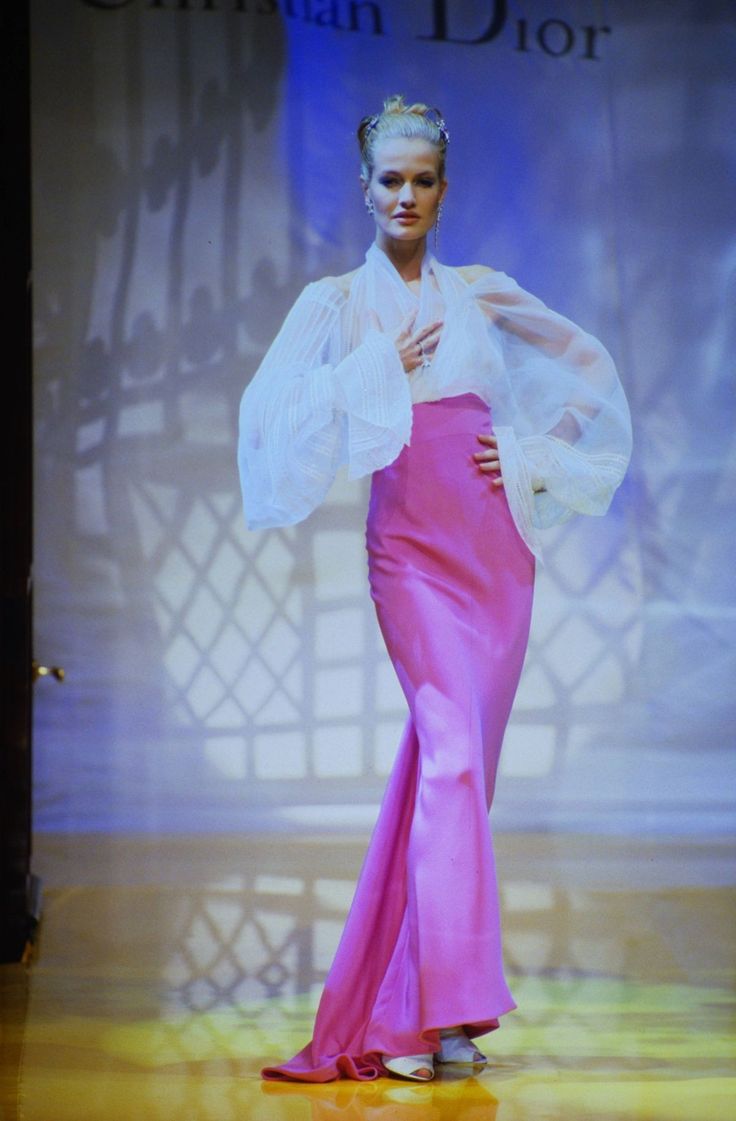 a runway model is posing on a fashion show stage, wearing a sheer white blouse and a long pink skirt. the background includes the christian dior logo and a windowlike design.