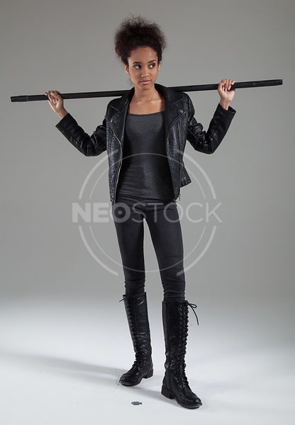 a young woman with black hair and dark eyes wearing a black leather jacket, black pants, and black boots is holding a black staff in front of her. she is standing on a grey background. she is looking away from the camera.