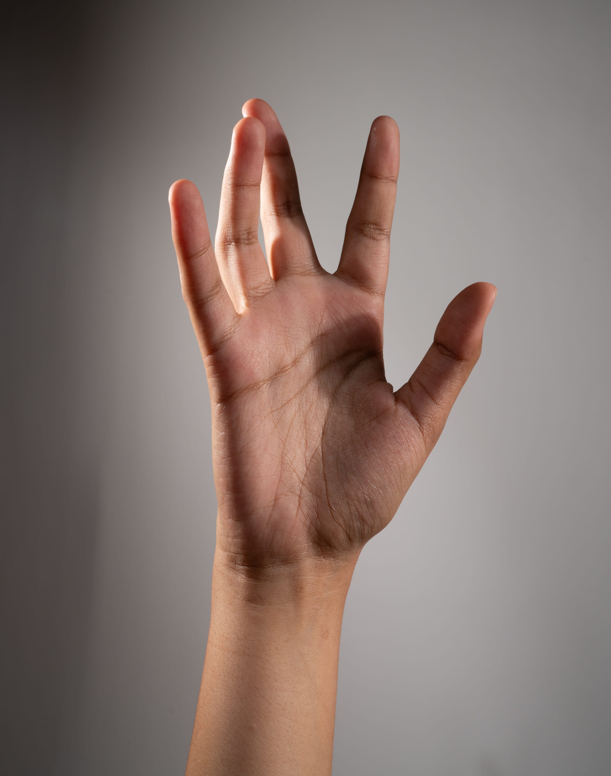 a single hand is held up against a neutral grey background, making the vulcan salute gesture from star trek. the hand is illuminated by a side light, which casts shadows and highlights the shape and texture of the skin.