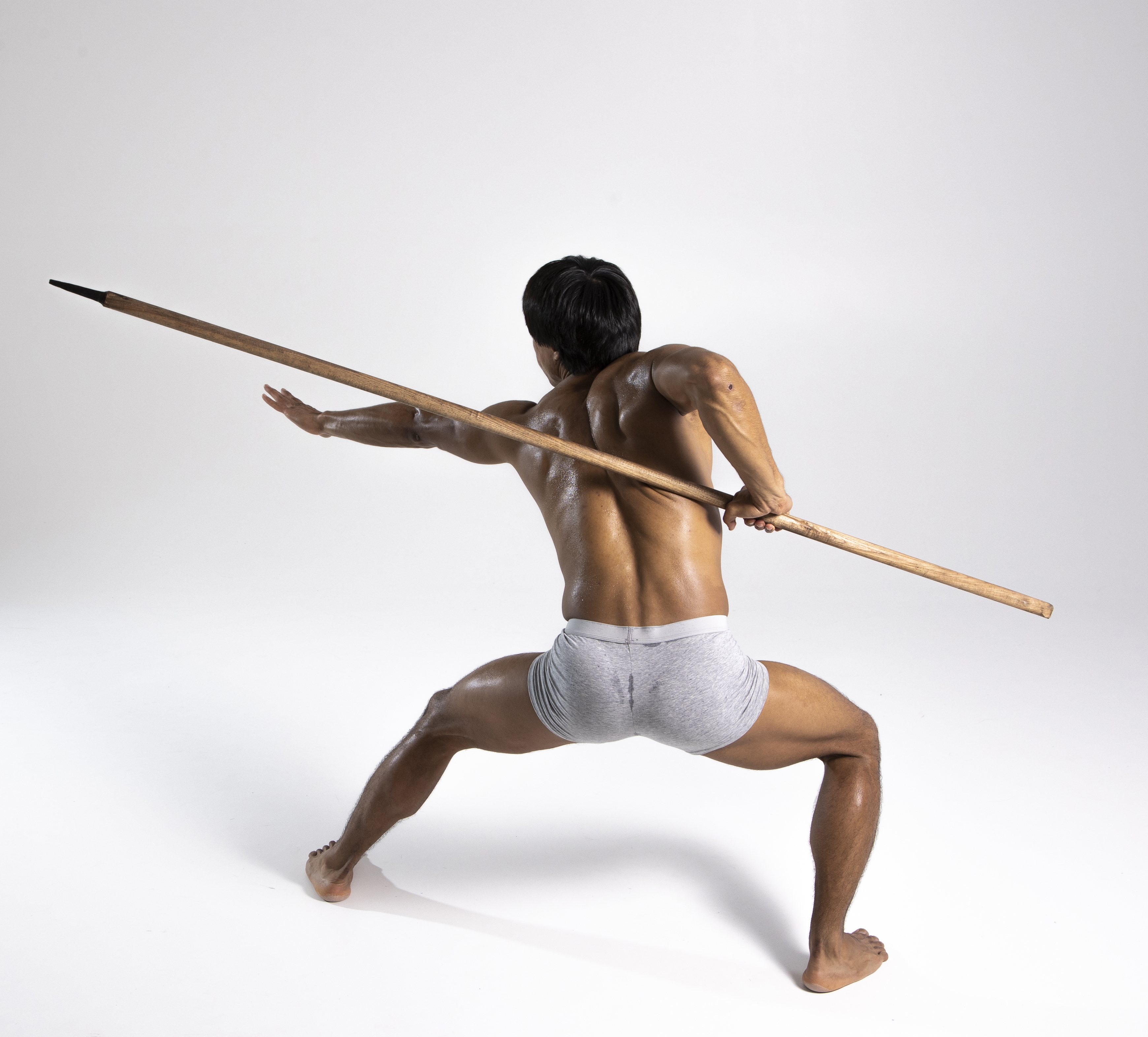 a muscular man is seen from the back, in a fighting stance, holding a spear. he is wearing gray briefs and is barefoot. the background is a plain white.