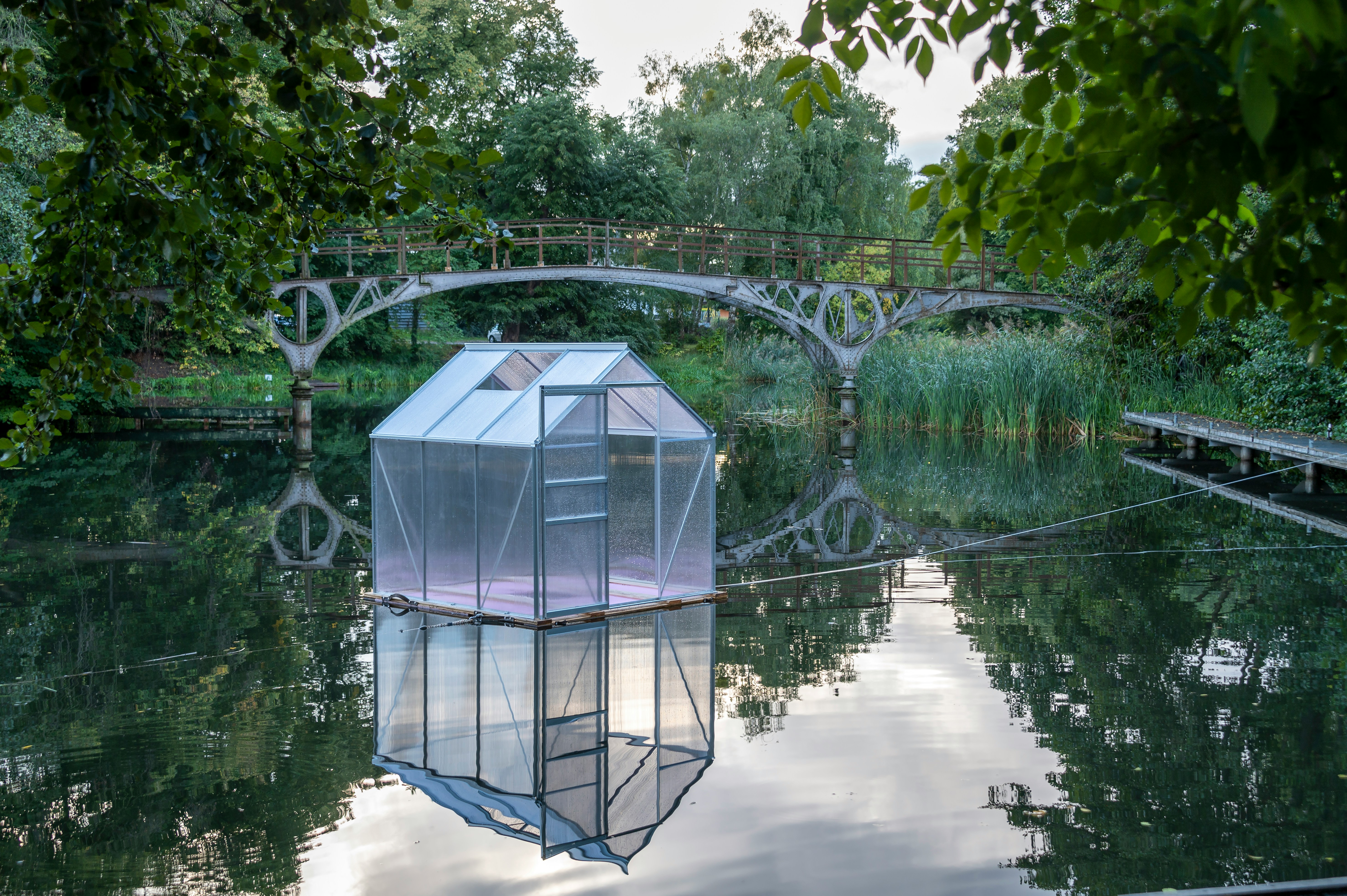 the image showcases a serene landscape featuring a small greenhouse floating on a calm body of water. a rustic bridge arches over the water in the background, connecting two banks adorned with lush greenery. the greenhouse's reflection is perfectly mirrored on the still water's surface, creating a captivating visual. the overall ambiance suggests a tranquil summer evening.