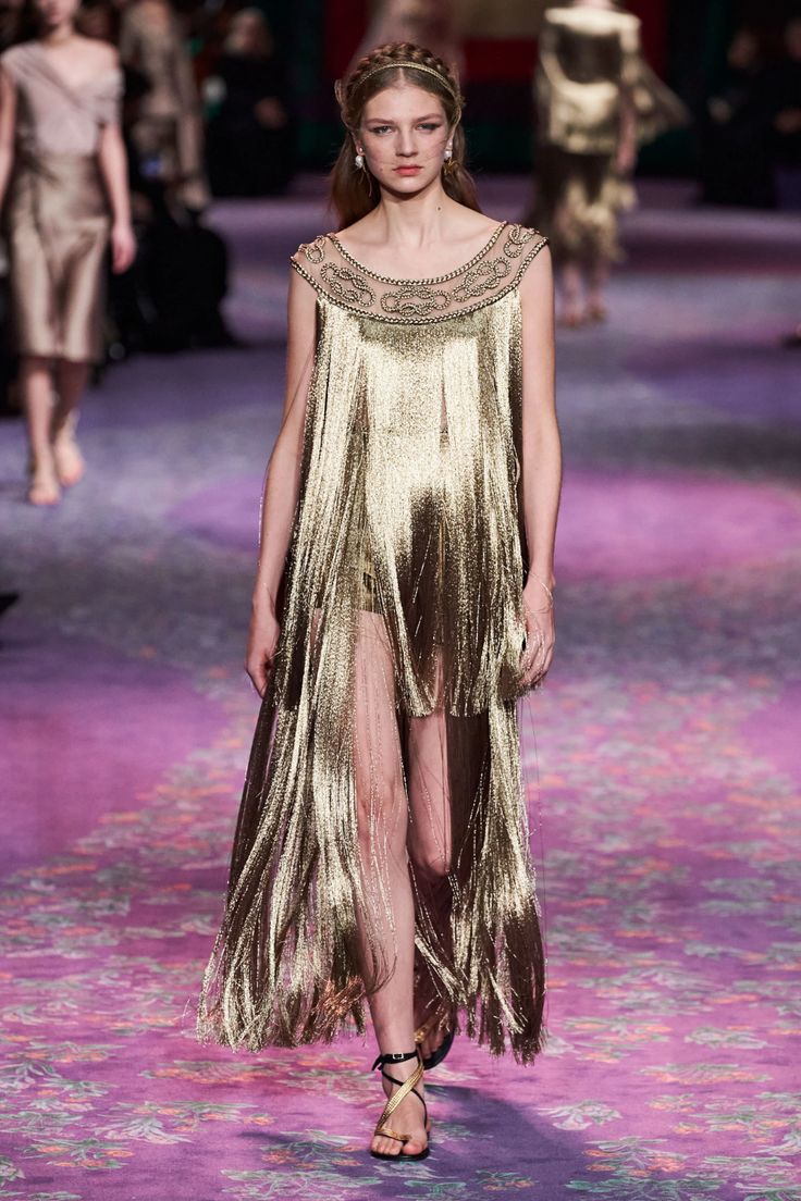 a fashion model walks down a runway in a gold fringe dress, her hair styled with a braid. the dress is adorned with embroidery and has a sheer panel at the bottom. she is wearing sandals with a gold strap. the runway is covered in a pink and purple floral pattern. the overall effect is elegant and glamorous.