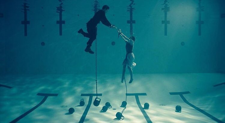 the image is a still from a film or a photograph styled like a film still. it depicts a man and a woman underwater in a swimming pool.  both are chained, with heavy weights attached to the chains, and are linked together by the chains. the man appears to be reaching towards the woman, but the overall tone is dark and suspenseful, implying a sense of captivity or tragedy. the scene is shot in a muted teal color palette, enhancing the atmospheric mood. the setting is a seemingly empty swimming pool, with lane lines visible on the bottom. the overall mood is dark, dramatic, and emotionally charged.
