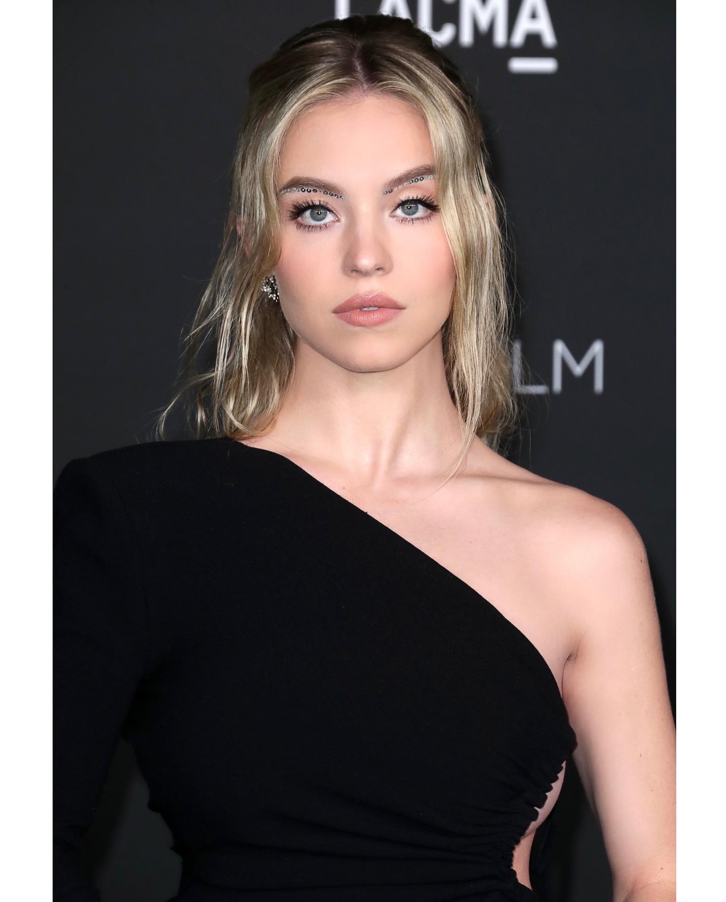 the image is a closeup portrait of a young woman, likely an actress, at what appears to be a formal event or red carpet. she's wearing a stylish black oneshoulder dress with long sleeves. her blonde hair is styled in loose waves, and she has on noticeable makeup, including a unique detail of glitter applied to her eyebrows. the background is dark and blurred, drawing attention to the subject. the overall impression is one of elegance and glamour.